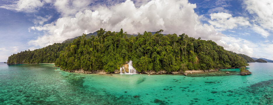 Aerial view of lush green island with cascading waterfalls meeting the turquoise sea under a partly cloudy sky, Togean Islands, Indonesia.