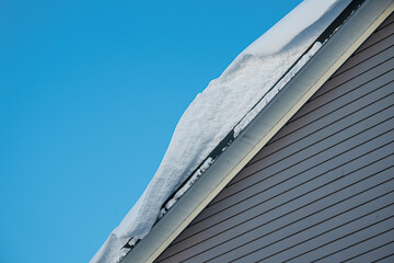 Heavy snow accumulation hanging from house roof edge against blue sky. © Trygve