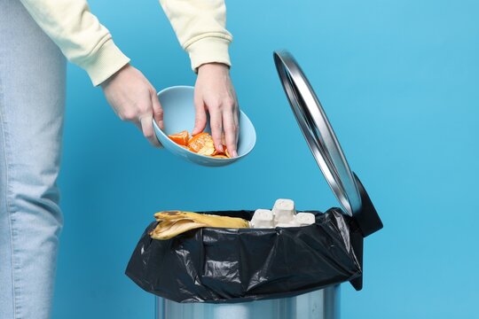 Woman throwing tangerine peels into trash bin on light blue background, closeup