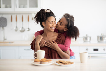 Cute black girl teenager hugging and kissing her happy beautiful mom after breakfast together,...