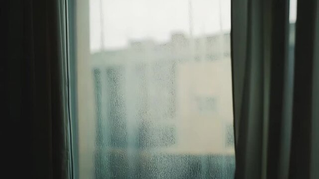Window pane streaked with rain droplets, view through curtains to a blurred urban building and wet street, moody gray atmosphere evoking solitude and quiet indoors