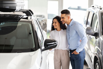 Happy middle-eastern young family choosing new auto in salon, standing by nice white vehicle,...