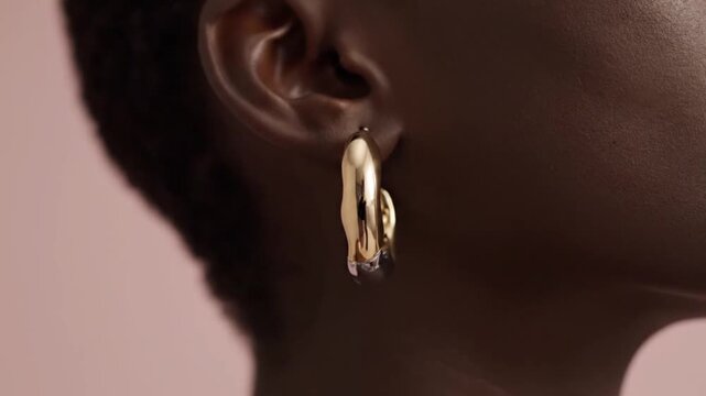 Close up profile view showcases a thick gold huggie hoop earring worn elegantly in the earlobe of a person with dark skin.