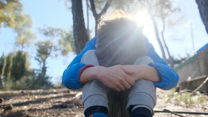 Young boy sitting alone on forest ground, embracing knees, contemplating deep thoughts in a moment of solitude and emotional struggle with sunlight flaring © DawDunia