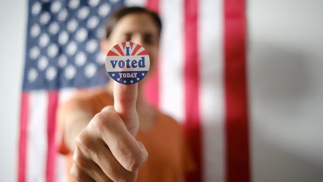 Person holding i voted today sticker on thumb with an american flag forming a patriotic background, representing civic duty, election, and democratic participation