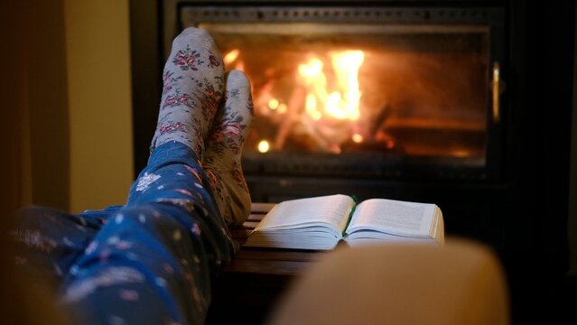 Person relaxing indoors by warm fireplace with feet up, wearing cozy socks and pajamas, reading a book during a cold winter evening, enjoying comfort and leisure at home