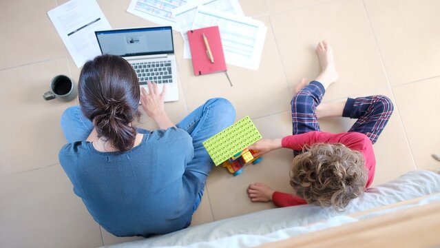 Woman focusing on work while sitting on the floor with laptop, documents, and coffee, managing a flexible career alongside a child playing with a toy car
