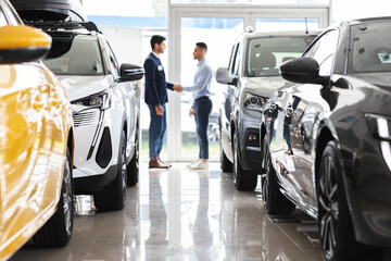 Middle-eastern young man customer and sales assistant shaking hands, car showroom interior, friendly manager greeting wealthy arab guy, standing among luxury brand new autos, full size shot