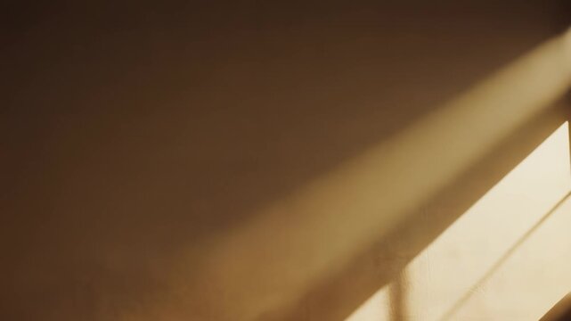 Warm sunlight streams onto an interior wall, casting sharp diagonal shadows and creating a beautiful abstract pattern of light and dark areas, suggesting a peaceful minimalist aesthetic