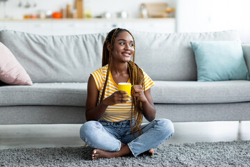 Fototapeta premium Happy black woman sitting on floor at home, drinking coffee, looking at copy space and smiling. Relaxed african american young lady in casual resting in living room, enjoying herbal tea