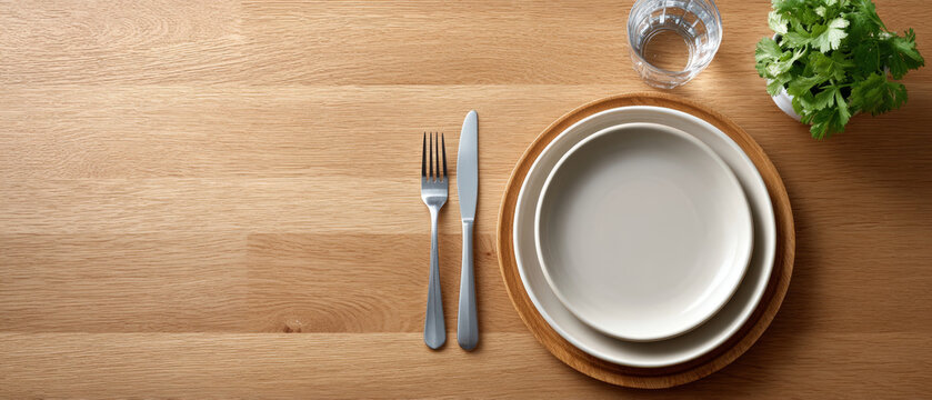 Minimalist dining table setting with empty white plates on wooden charger, stainless steel fork and knife, glass of water, and fresh green herbs on natural wood surface