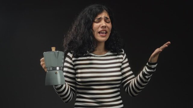Young brunette woman in striped top holding a moka pot and shrugging with palms up in black studio; coffee frustration.