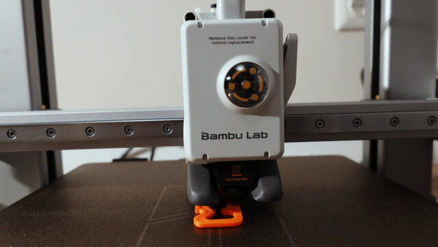 Close-up of a 3D printer nozzle depositing an orange plastic object on a build plate