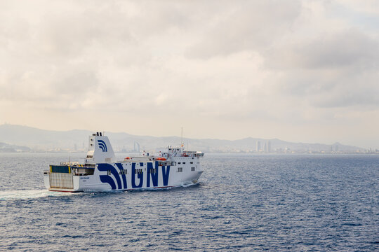 PORT OF BARCELONA, SPAIN;A - JULY 2, 2024 A large white GNV ferry sails through the calm sea, leaving a wake, with a coastal city and mountains visible in the background under a cloudy sky.