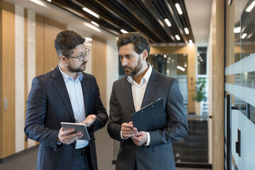 Two businessmen walk through a modern office corridor, sharing ideas from a tablet and clipboard...