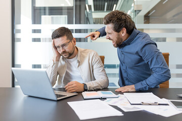 Aggressive executive is shouting and pointing at a frustrated male employee working on a laptop,...