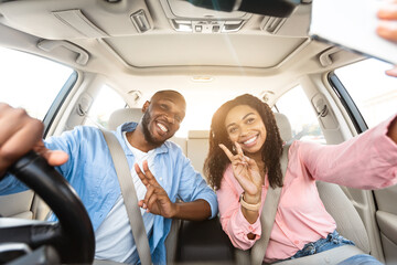 Happy black lovers taking selfie while having car trip, sitting in auto, cheerfully smiling at...