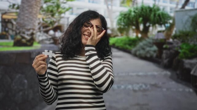 Young arab brunette woman holds a puzzle piece and covers her face with hand on a street near green park trees; amused curiosity.