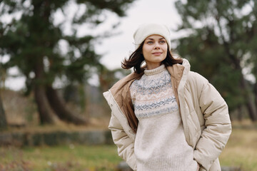 Woman in a warm coat and knitted sweater stands outdoors in a park during chilly weather, wearing a...