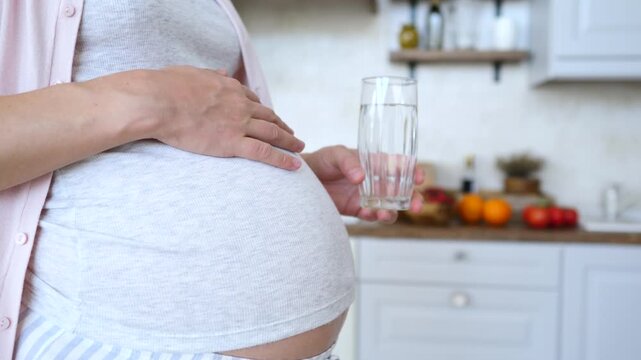 Expectant mother holding a glass of pure water while gently stroking her tummy in the kitchen, promoting a healthy lifestyle, hydration, and well being during her pregnancy journey