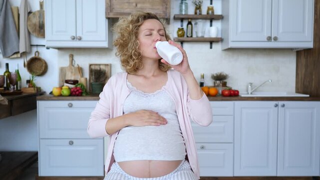 Pregnant woman with a bare belly feeling unwell from heartburn symptoms, drinking milk or yogurt from a bottle to find relief from indigestion and stomach pain while sitting in her kitchen