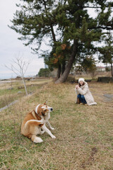 Obraz premium A woman kneels beside a dog in a grassy park, photographing the canine with a camera, capturing a candid outdoor moment amid trees and open landscape