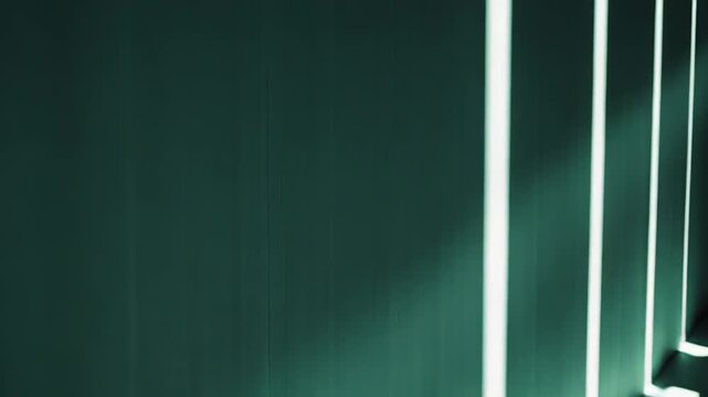 Sunlight streams through an unseen window, casting strong, parallel lines of light and shadow across a teal wall and concrete floor, creating a minimalist, atmospheric scene