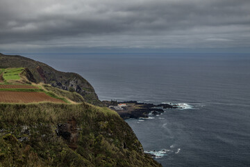 A high-angle view of the dramatic volcanic coastline at Ponta da Ferraria. Rugged cliffs and agricultural fields overlook the natural thermal baths and the Atlantic Ocean in the Azores.