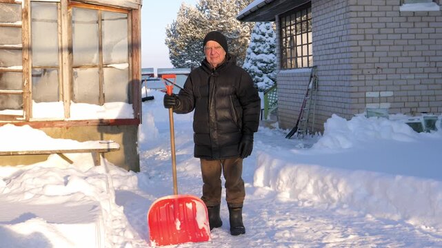 Elderly man in warm winter clothes standing in backyard with snow shovel. Looking at camera after clearing path from the heavy snowfall during sunny cold day. Person clearing snow outdoors.