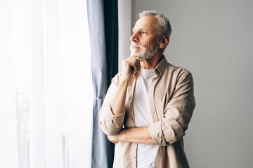 Happy positive senior man wearing casual clothes standing near window looking away