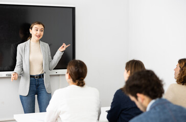 Fototapeta premium Young female business coach explaining topic while standing against group of people sitting at desks in front of her
