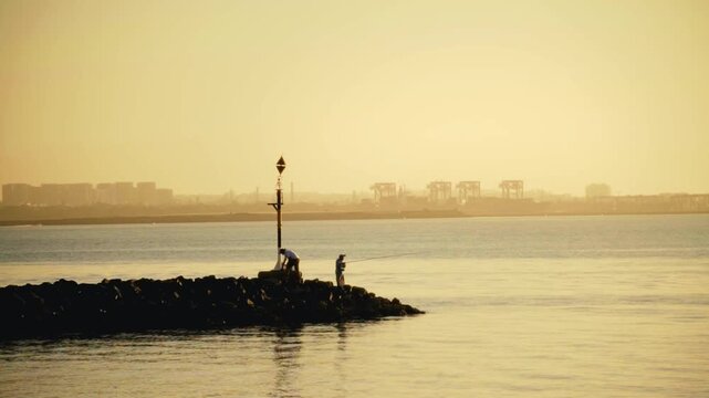 Fishermen on Rocky Breakwater at Ramsgate Beach During Golden Sunrise with Industrial Port Skyline in Background
