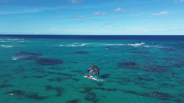 Aerial footage over open sea following a parasailing boat towing a colorful parachute