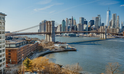 Fototapeta premium Brooklyn Bridge and Manhattan skyline during a clear day in New York City. The Brooklyn Bridge stands tall as boats pass under it.