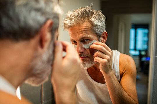 Mature man applying under-eye patches in bathroom mirror