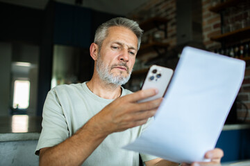 Mature man checking bills on smartphone at home kitchen