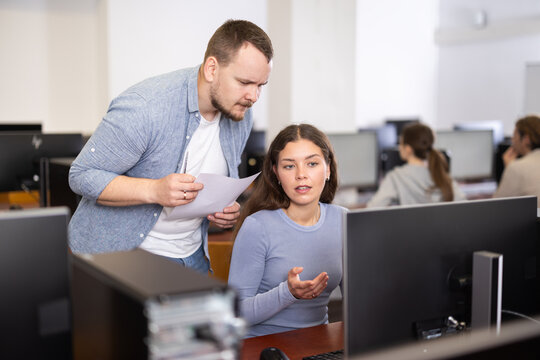Male teacher of computer courses helps girl student to figure out with performance of difficult task, master ark. Mentor observes students studies, looks at monitor screen.