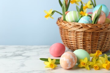 Painted Easter eggs and beautiful daffodils in wicker basket on white marble table against light blue background, closeup. Space for text