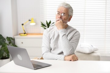 Fototapeta premium Amnesia. Senior man with laptop trying to remember something at white table indoors