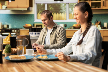 Fototapeta premium Busy working dad typing on laptop at kitchen table with his family, ignoring his pre-teen girl and wife during breakfast. Distracted preoccupied father distant with his daughter.