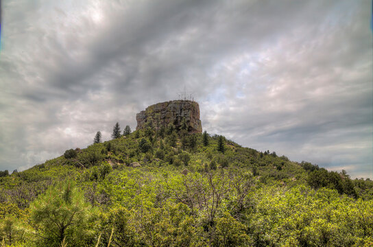 Castle Rock Colorado. A rocky cliff rises above a lush, green landscape under a partly cloudy sky. Tall trees are scattered around the hill, and the scene is brightened by the midday sun.