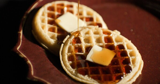 Maple syrup being poured over two toasted frozen waffles with pats of butter on a rustic brown ceramic plate