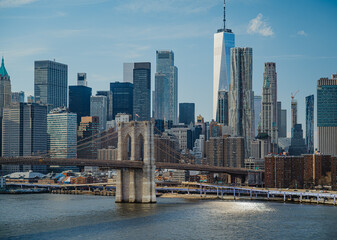 Fototapeta premium City skyline view with bridges and buildings under blue sky in New York. Tall buildings stand close to the water with a bridge connecting parts of the city on a bright day.
