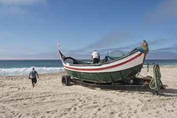 Fototapeta premium Fishing scene on the beach in Leirosa, Portugal, featuring a traditional boat with a curved prow and stern painted in green, white, and red. The boat rests on a trailer 