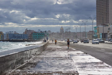 Obraz premium Panoramic view of Havana’s Malecon on a cloudy day, with rough sea waves crashing against the seawall. In the background, the urban skyline and iconic architecture of Havana facing the ocean.