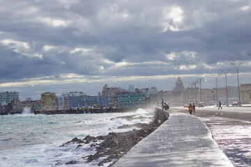 Obraz premium Panoramic view of Havana’s Malecon on a cloudy day, with rough sea waves crashing against the seawall. In the background, the urban skyline and iconic architecture of Havana facing the ocean.