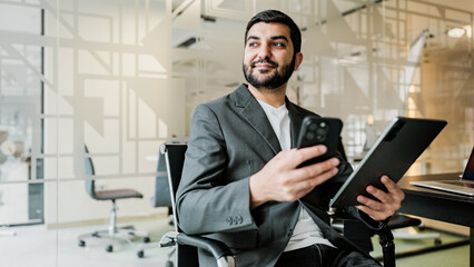 Young man sits in modern office and uses smartphone while looking ahead with a focused expression during work hours