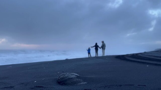 Static shot of black sand beach with small rock in foreground and distant people walking under cloudy sky, Iceland. HD 1080p.
