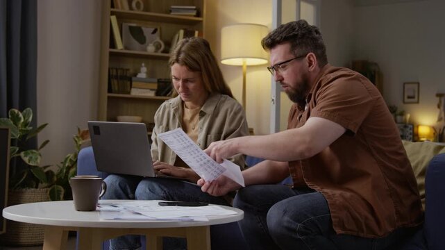 Medium shot of Caucasian adult couple sorting financial documents and using laptop while managing home finances together, copy space