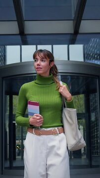 Vertical. Tracking shot of a caucasian millennial woman in a green sweater walking out of a modern office entrance as coworkers pass by, conveying confidence, career momentum and urban corporate life.